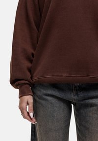 Felpa oversize marrone con polsini a costine, abbinata a jeans grigi a vita alta. Tessuto morbido, design casual e vestibilità rilassata.