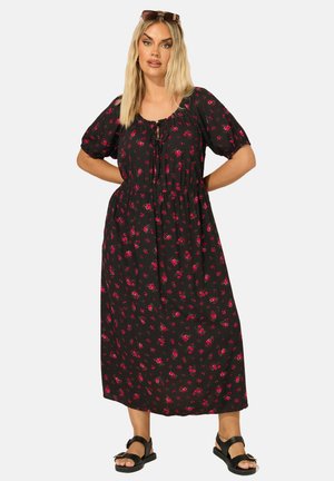 Donna con capelli biondi lunghi indossa un vestito midi nero con piccola stampa floreale rossa, maniche a sbuffo, scollo annodato, sandali neri e occhiali da sole sulla testa.