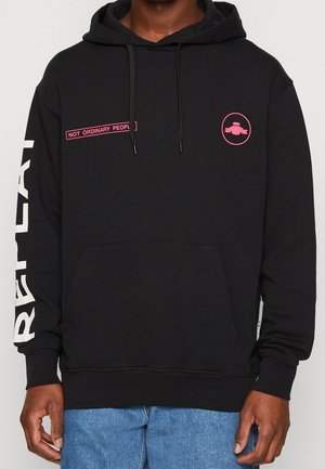 Czarna bluza z kapturem z różowym napisem "NOT ORDINARY PEOPLE" na klatce piersiowej, okrągłym różowym logo oraz dużym białym napisem na lewym rękawie, noszona z niebieskimi dżinsami.