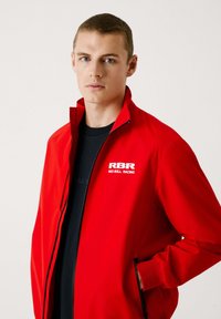 Rode ritsjack in een levendige rode kleur, voorzien van zwarte ritszakken, zacht materiaal en een wit logo op het linkerborstgedeelte.