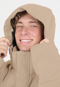 Beige isolerad jacka med huva, med en slät yta, justerbara dragsko och hög krage. Enkelt design och minimala accenter.