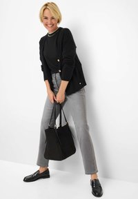 Cardigan nero con bottoni dorati, camicia nera, jeans grigi e mocassini neri. Tiene una borsa a tracolla nera strutturata. Texture liscia.