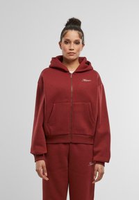 Bordový zipsový mikina s priestrannou kapucňou, rebrovanými manžetami a prednými vreckami. Obsahuje malé logo na hrudi a mäkkú, textúrovanú tkaninu.