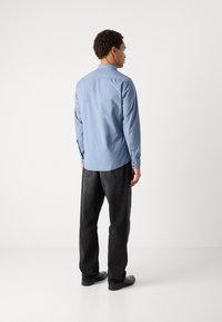 Long-sleeve light blue shirt with a button-down collar, featuring back yoke detail and curved hem, paired with dark jeans and black shoes.