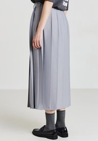 Jupe plissée grise avec une texture lisse, longueur mi-mollet et fentes sur les côtés. Associée à des chaussures noires et des chaussettes grises.