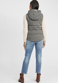 Graue Steppweste mit Kapuze, ausgestattet mit einem gesteppten Design und beigen Ärmeln, kombiniert mit hellblauen Jeans und braunen Stiefeletten.