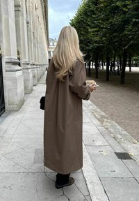 Ξανθιά γυναίκα με μακρύ καφέ παλτό κρατάει φαγητό, περπατώντας σε λιθόστρωτο μονοπάτι δίπλα σε κτίριο με κολόνες και περιοχή πάρκου με δέντρα.