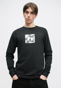 Νέος με κοντά καστανά μαλλιά φοράει μαύρη μπλούζα με μακριά μανίκια και λευκό στιλιζαρισμένο λογότυπο "h" και αστέρι, με τα χέρια στις τσέπες μαύρων παντελονιών.