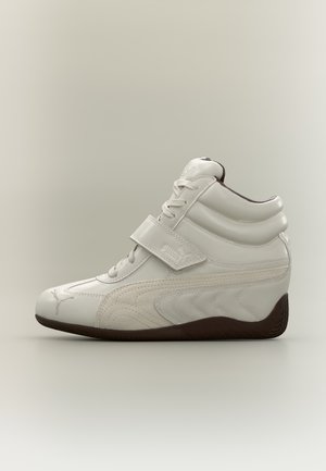 Basket montante Puma blanc avec des lacets, une bride à la cheville, des empiècements latéraux cousus et une semelle marron foncé, sur un fond uni.