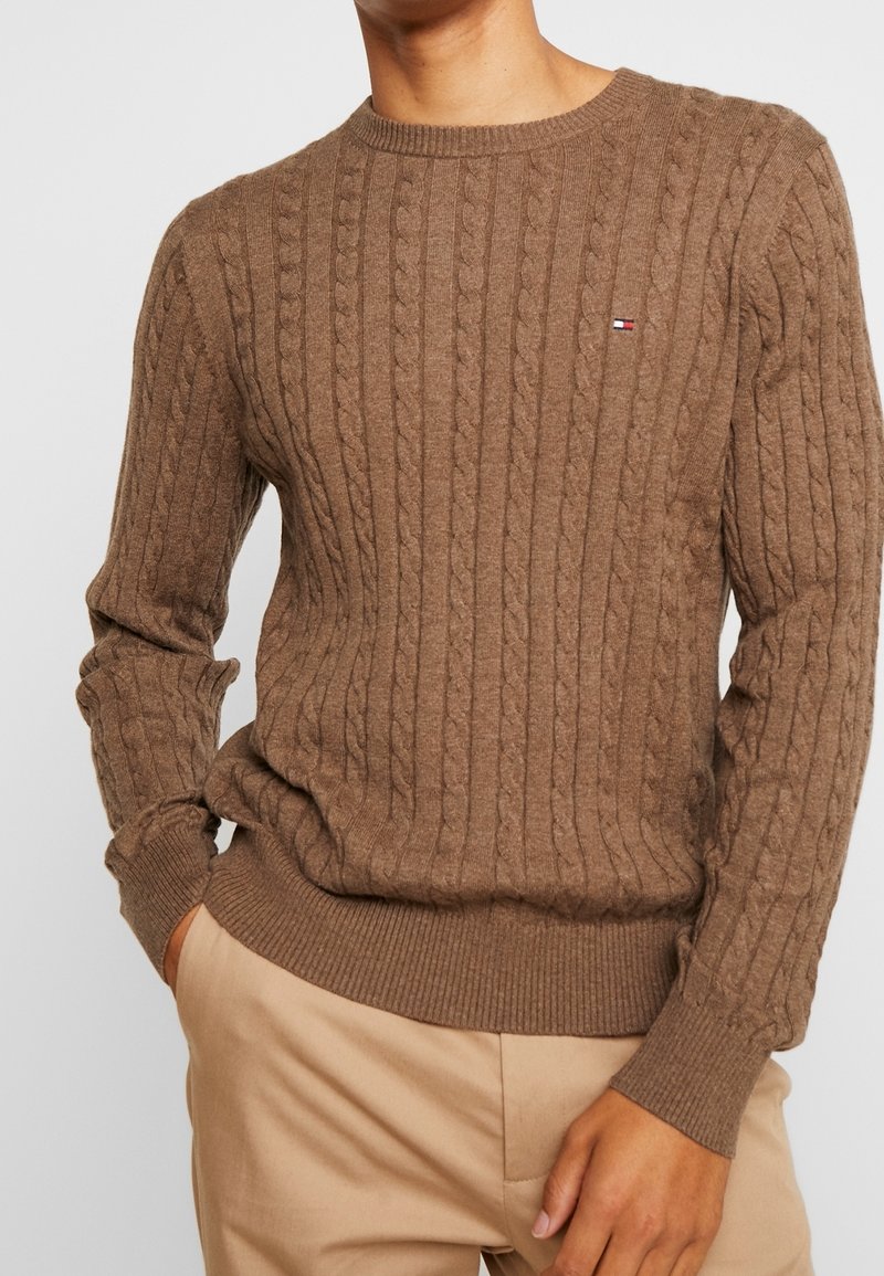 Pull en tricot marron avec une texture côtelée et un motif en torsade. Il possède un col rond et un petit logo sur la poitrine.