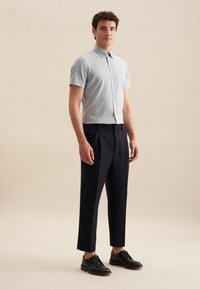 Camicia azzurra a maniche corte con bottoni, abbinata a pantaloni blu scuro e scarpe nere con lacci, in piedi davanti a uno sfondo neutro.