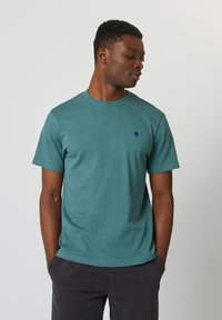 Teal katoenen t-shirt met een subtiele gespikkelde textuur, ronde halslijn, korte mouwen en een klein donkerblauw boomlogo op de borst.