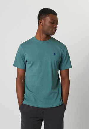 Teal katoenen t-shirt met een subtiele gespikkelde textuur, ronde halslijn, korte mouwen en een klein donkerblauw boomlogo op de borst.