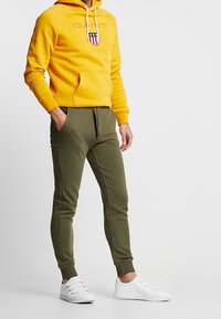 Sudadera con capucha amarilla con bolsillo frontal y logo, combinada con pantalones jogger verde oliva y zapatillas blancas. Diseño de corte casual.