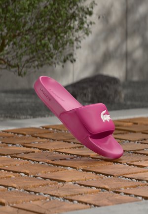 Sandalo rosa senza lacci con una texture liscia, caratterizzato da una fascia larga e dal logo Lacoste. Posato su piastrelle di legno con uno sfondo naturale.