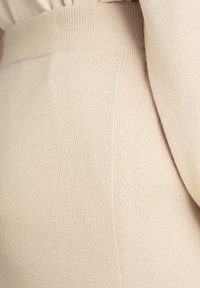 Tissu tricoté beige avec une texture côtelée. Le matériau est doux avec une finition lisse, présentant des lignes verticales discrètes tout au long.