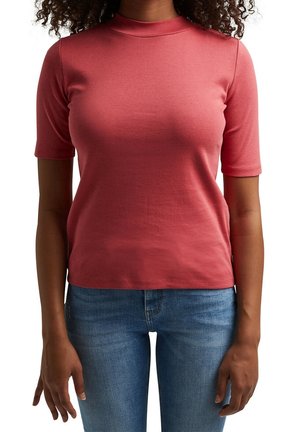 Nainen, joka on pukeutunut tiukkaan vaaleanpunaiseen lyhythihaiseen Collegepaitaan ja sinisiin farkkuihin, seisoo valkoista taustaa vasten, kädet rentoina sivuilla.