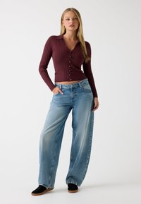 Bordeauxrotes, geripptes langärmliges Oberteil mit Kragen und Knöpfen, kombiniert mit hellblauen, weiten Jeans und schwarzen Sneakers mit Gummisohlen.