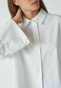 Chemise blanche en coton à boutons avec un col pointu et des poignets larges, présentant une texture lisse et un design épuré, mettant l'accent sur la simplicité.