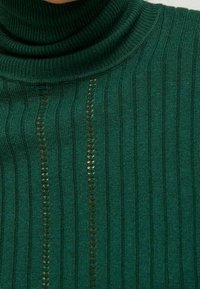 Grön ribbad tröja med hög krage, med vertikala ränder och dekorativa öppna stickade sektioner längs fronten. Mjuk textur.