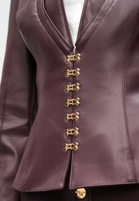 Detailný záber na bordovú koženú bundu so šiestimi zlatými háčikmi a očkami po celej prednej strane, nosenú cez biely top.