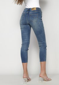 Jeans de mezclilla azul de corte medio y cropped con un acabado deslavado. Cuenta con dos bolsillos traseros y dobladillo deshilachado. Combinados con sandalias de tacón de bloque transparentes.