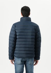 Navy blue padded jacket with a high collar, featuring horizontal quilted sections, and a smooth, shiny texture, worn with denim jeans.