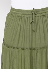 Hame olive green dengan tekstur berkerut, pinggang elastis, dan penutupan tali. Memiliki detail tepian berenda.
