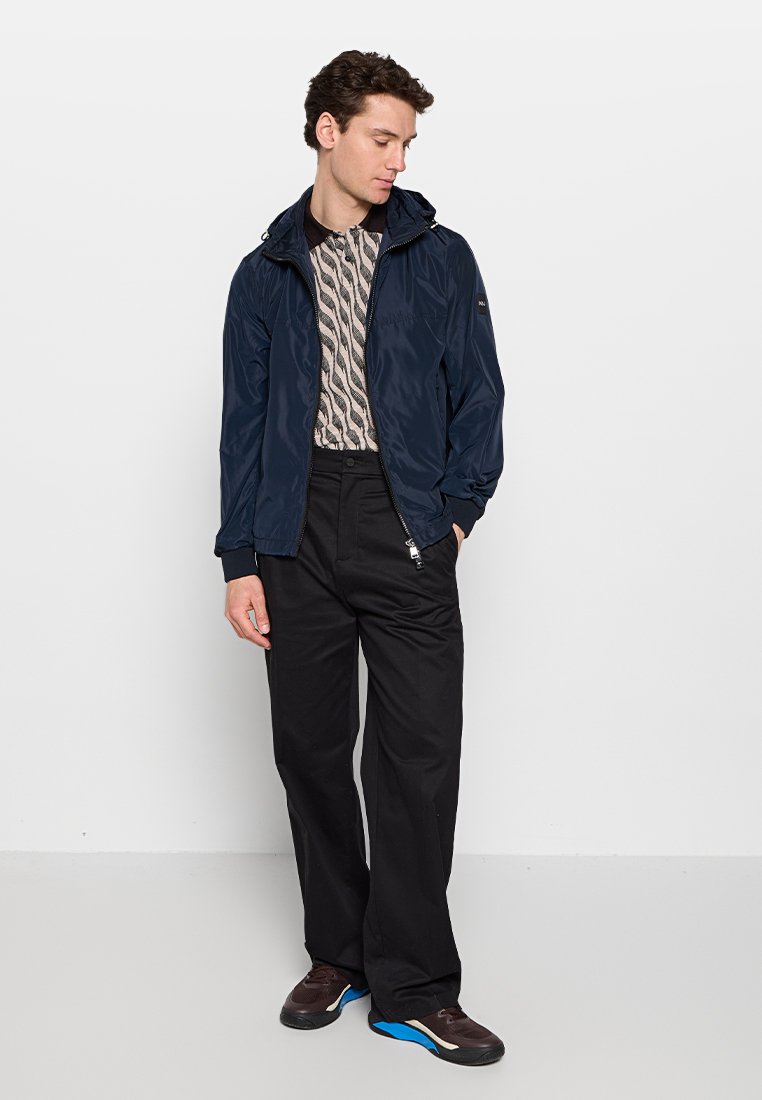 Dunkelblau, leichtes Jackett mit Reißverschluss und Kapuze; getragen über einem gemusterten Hemd. Kombiniert mit schwarzen Hosen und dunklen Schuhen.