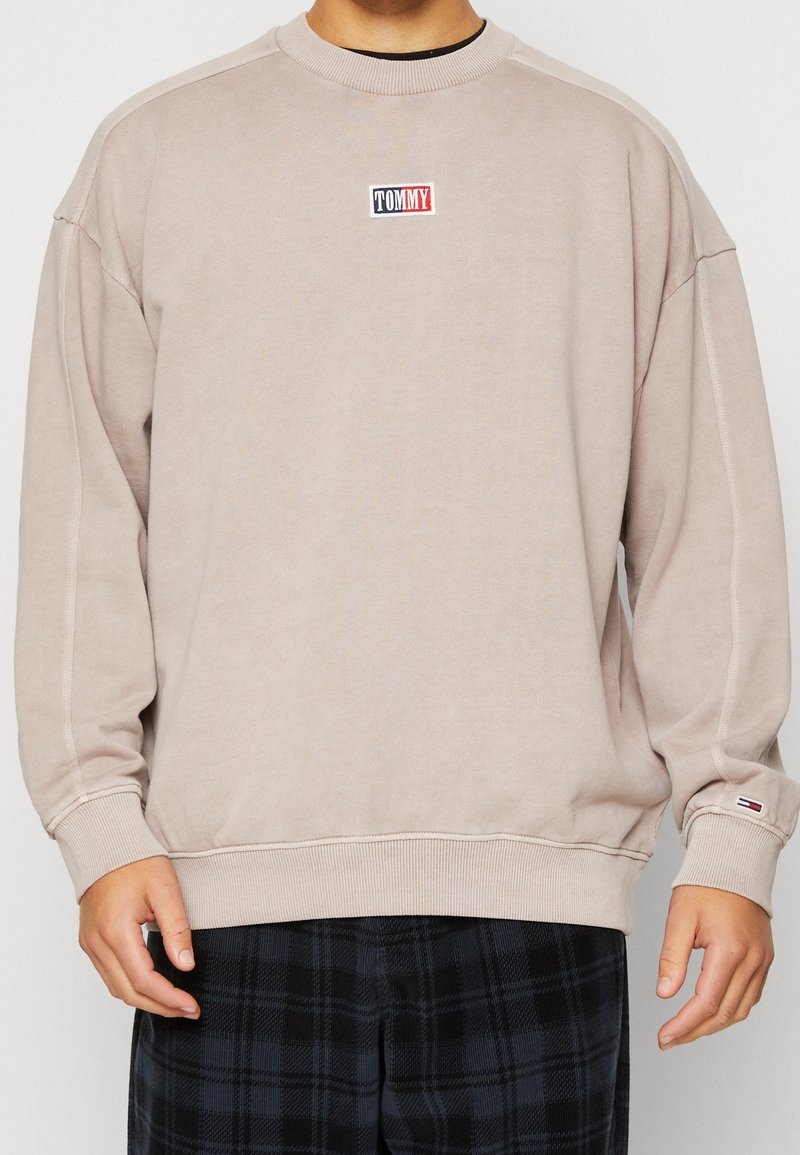 Sudadera color beige claro con cuello redondo, puños y dobladillo acanalados. Presenta un pequeño logotipo rojo, blanco y azul en el pecho. Ajuste suave y relajado.