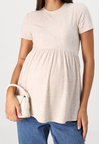 Kortärmad, räfflad amningstopp i ljusbeige med utsvängd nederkant. I kombination med en liten, krämfärgad handväska. Blå denimjeans synliga.