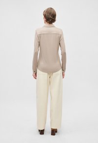 Bež košulja s dugim rukavima i zaobljenim rubom, s detaljima na leđima, u kombinaciji s kremasto bijelim širokim hlačama i smeškim cipelama na blok petu.