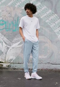 Hvid kortærmet t-shirt med et sort logo, lyseblå træningsbukser og hvide sneakers med pastelfarvede accenter, sat mod en graffiti-baggrund.
