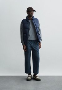 Jūrinis pūkuotas liemenė, pilkas džemperis, laisvi mėlyni kelnių, rudas kepurė, ir daugiaspalviai batai su kontrastiniu padu, ant neutralios fono.