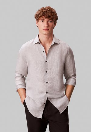 Ung mann med krøllete hår som har på seg en lysegrå skjorte med knapper, med oppbrettede ermer og svarte bukser, stående med hendene i lommene.