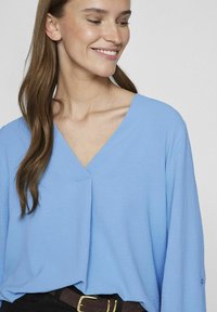 Maglia a maniche lunghe di un blu chiaro con scollatura a V e vestibilità rilassata. Tessuto testurizzato si adagia dolcemente sulle spalle. Indossata con una cintura scura.