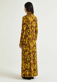 Langes gelbes Kleid mit braunem Blumenmuster. Es hat einen hohen Kragen und lange Ärmel und besteht aus einem leichten, strukturierten Stoff.