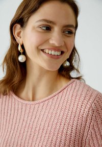 Helles rosa gestricktes Sweater mit gerippter Textur und rundem Halsausschnitt. Das Modell trägt goldene Tropfenohringe mit großen weißen Perlen.