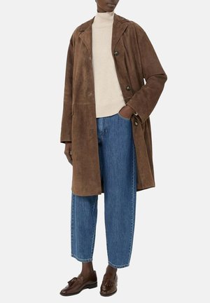 Persona che indossa un cappotto in camoscio marrone fino al ginocchio, un dolcevita crema, jeans blu e mocassini marroni con nappine, in piedi con una mano in tasca.