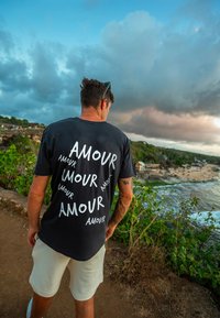 Czarny bawełniany t-shirt z dużym białym napisem "AMOUR" w różnych orientacjach. Noszony z jasnymi szortami. Tło zawiera zieleń i krajobraz morski.