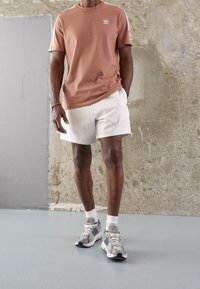 Ljusbrun kortärmad T-shirt med vita ränder, kombinerad med vita shorts och grå sneakers med svarta detaljer och ljusa snörningar.