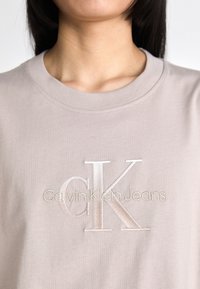 Šviesiai smėlinės spalvos marškinėliai su apvaliu iškirpimu, puoštas išsiuvinėtu logotipu: "Calvin Klein Jeans" blizgiu pabaigu. Minkšta medvilnė.
