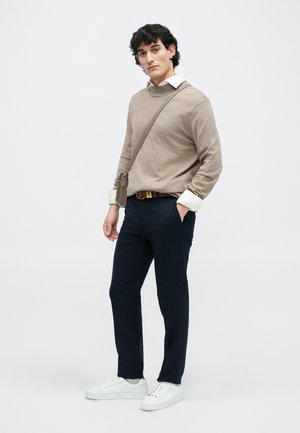 Suéter beige texturizado, collar blanco, pantalones azul marino y zapatillas blancas. Cinturón marrón con hebilla dorada, bolso bandolera en tono suave.