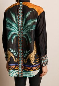 Chemise noire à manches longues avec motifs colorés de palmiers et léopards, accents géométriques, et texture de tissu lisse, avec des boutons décoratifs sur les manches.