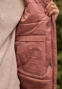 Padded jacket w miękkim różowym materiale, z gładką teksturą, etykietami wodoodpornymi i wiatroszczelnymi, kieszenią oraz zapięciem na napy wewnątrz.