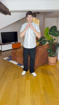 Bílá košile s vertikálními pruhy a krátkým rukávem, černé kalhoty a bílé boty. Stojí na dřevěné podlaze v příležitostném interiéru se rostlinami.