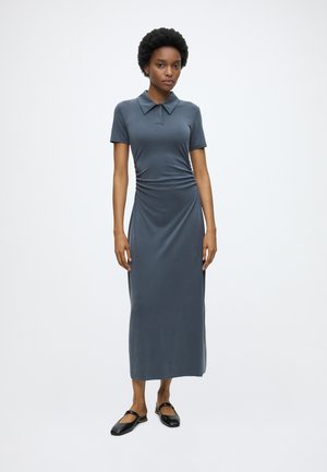 Woman standing wearing a short-sleeve, collared, ankle-length gray dress and black flat shoes on a white background.