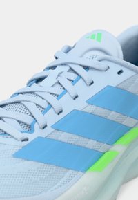 Αθλητικό παπούτσι ανοιχτού γαλάζιου χρώματος με επάνω μέρος από δίχτυ, μπλε λεπτομέρειες, πράσινη λεπτομέρεια στη φτέρνα και λευκούς κορδονιούς. Το λογότυπο της Adidas είναι ορατό στη γλώσσα.