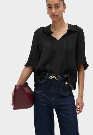 Kvinde iført sort bluse med flæsekant, stoppet ned i mørkeblå jeans med et bælte med guldlås, holder en bordeaux håndtaske mod en hvid baggrund.