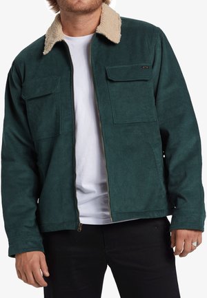 Chaqueta de pana verde azulado con collar de piel sintética beige, cierre de cremallera, dos bolsillos en el pecho y un corte ligeramente corto. Llevada sobre una camiseta blanca.